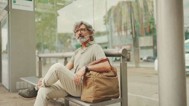 Thoughtful, middle-aged man with gray hair and beard, wearing casual clothes, sits at public transport stop. Mature gentleman in eyeglasses with bag is waiting for the tram at the tram station - Valokuva, kuva