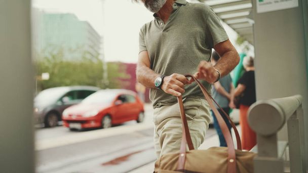 Middle-aged man with gray hair and beard, wearing casual clothes, gets on tram at public transport stop. Mature gentleman in eyeglasses with bag on his shoulder gets on the tram - Photo, image