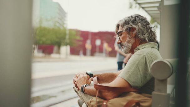 Thoughtful, middle-aged man with gray hair and beard, wearing casual clothes, sits at public transport stop. Mature gentleman in eyeglasses with bag is waiting for the tram at the tram station - Фото, изображение