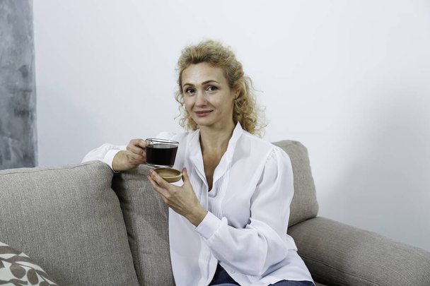 Portrait of adult beautiful woman wearing a white blouse sitting on the couch holding a big glass cup of black coffee. Copy space, background, close up - Фото, изображение