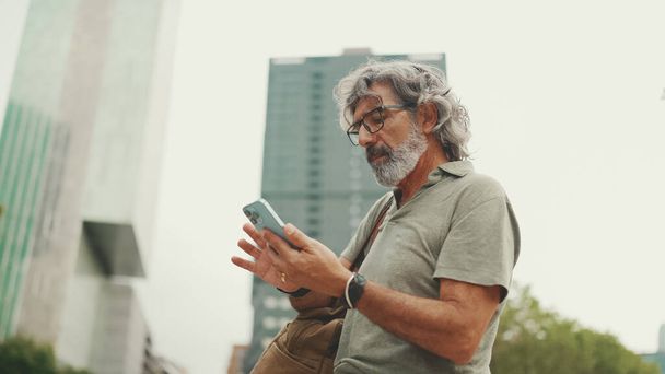 Friendly middle aged man with gray hair and beard looking at map trying to find his way using his mobile phone. Mature gentleman in eyeglasses using map app in cellphone outdoors - Φωτογραφία, εικόνα