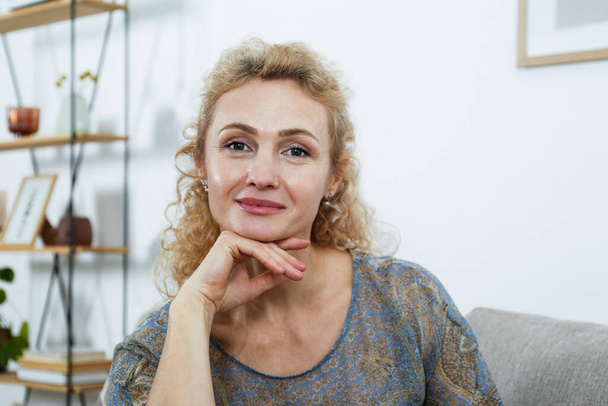 Portrait of adult beautiful woman with curly blond hair sitting on the couch and smiling. Close up shot of happy middle aged female. Copy space, background. - Photo, Image