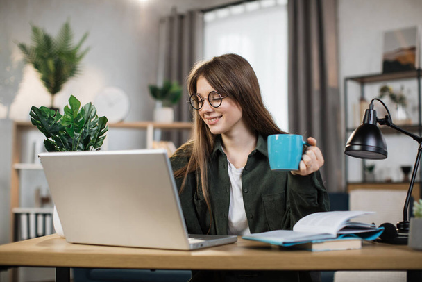 Attractive female freelancer with cup of coffee in casual outfit using modern laptop for work at home. Caucasian woman with dark hair sitting at table and typing on computer. - Foto, imagen