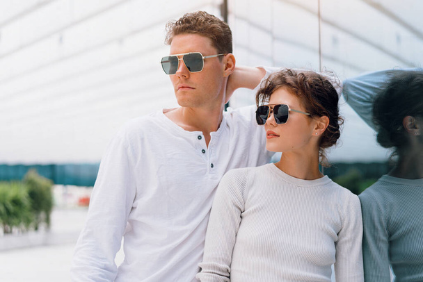 Smiling beautiful woman and her handsome boyfriend. Woman in casual summer jeans. Serious pensive couple in sunglasses walking business district. Couple posing on the street modern building background - Foto, imagen
