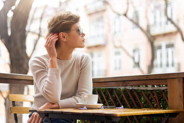 Portrait of thoughtful young woman sitting at table with coffee and looking away, pensive charming young woman posing at free time and waiting friend or boyfriend. - Foto, Bild