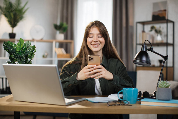 Confident young woman writing message during mobile conversation at office. Caucasian female worker with long dark hair sitting at table with opened laptop. - Foto, afbeelding