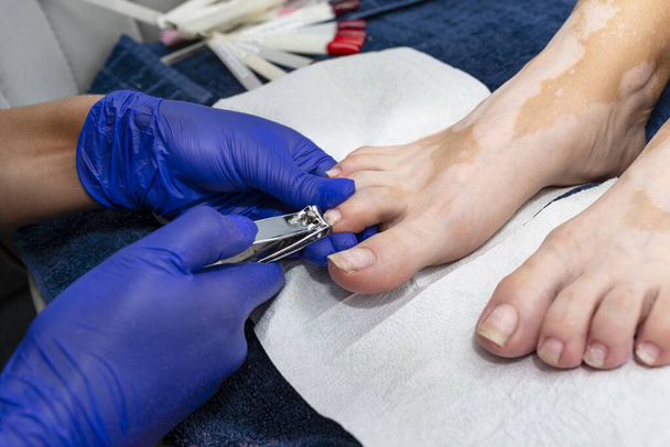 The manicurist cuts her toenails with a nail clipper wearing blue latex gloves. - Fotó, kép