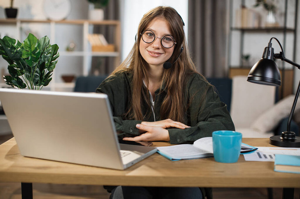 Young caucasian woman in headset sitting at desk with laptop and talking during video call. Businesswoman using modern devices for online conversation at work. - Фото, изображение