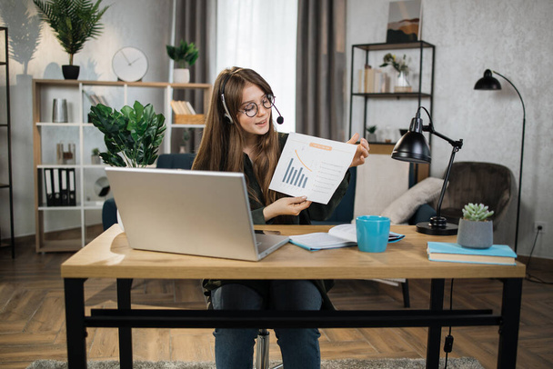 Caucasian woman presenting financial report to colleagues through video call on laptop. Female freelancer sitting at workplace and talking in headset. - Foto, imagen