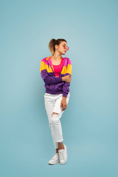 Full length of self assured young female millennial with ponytail in stylish outfit and sunglasses looking away while standing against blue background - Foto, immagini
