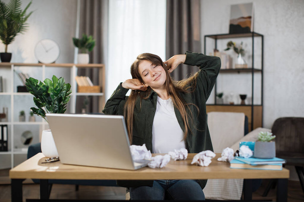 Gorgeous office woman relaxing by stretching her body while sitting in front her computer laptop at the wooden working desk over comfortable living room as background. - Fotografie, Obrázek