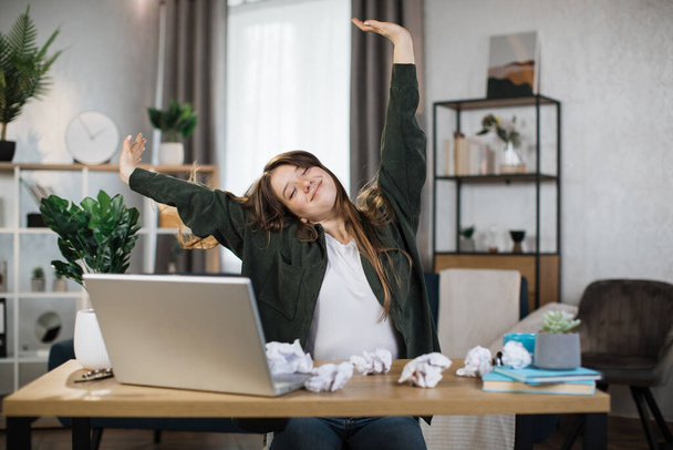 Gorgeous office woman relaxing by stretching her body while sitting in front her computer laptop at the wooden working desk over comfortable living room as background. - Foto, afbeelding
