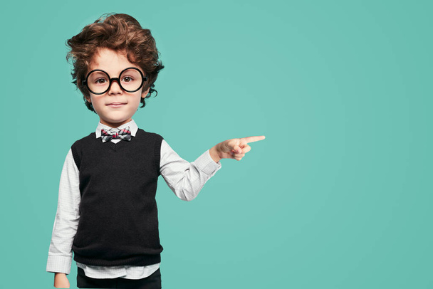 Adorable smart little boy with dark hair and big head in formal outfit and round eyeglasses, pointing aside and looking at camera against turquoise background - Foto, Imagen