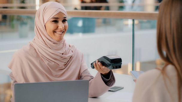 Friendly muslim woman in hijab seller agent offers pay for service through bank terminal girl shopper consumer pays for purchase using contactless payment technology on smartphone client uses phone - Фото, изображение