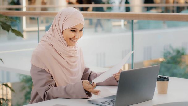 Genç İslami kadın kafeteryada oturmuş video konferansında sohbet ediyor. Laptop mesafesindeki tesettürlü müslüman kız internet kamerasındaki uzaktan görüşmede müşteriyle pazarlık yapıyor. - Fotoğraf, Görsel
