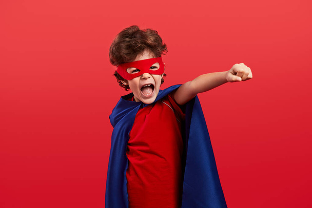 Excited little boy in cape and mask clenching fist and yelling while pretending to be superhero against red background - Zdjęcie, obraz