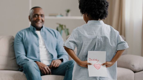 African american family mature dad sitting at home on couch surprised accepting birthday greetings little loving daughter gives handmade card for fathers day child comes to happy man hugs tightly - Foto, imagen
