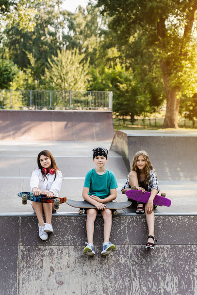 Children friends company with skateboard and penny boards on skate board park. Extreme lifestyle. Active kids sitting on sport ramp, communicate with each other and have fun - Foto, immagini