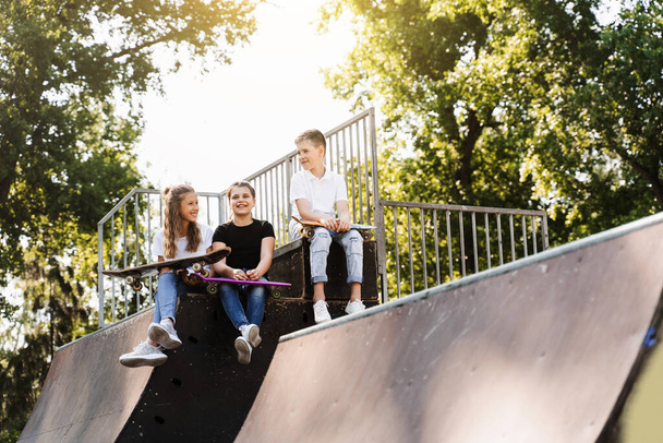 Спортивные дети со скейтбордом и пенни-бордами сидят и болтают друг с другом на спортивной рампе на детской площадке. Детское общение и дружба. Экстремальный образ жизни - Фото, изображение