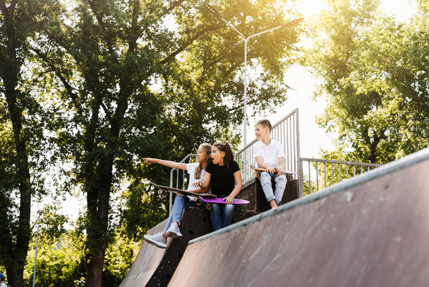 Sports kids with skateboard and penny boards are sitting and chatting with each other on sports ramp on playground. Children communication and friendship. Sports extreme lifestyle - Fotoğraf, Görsel