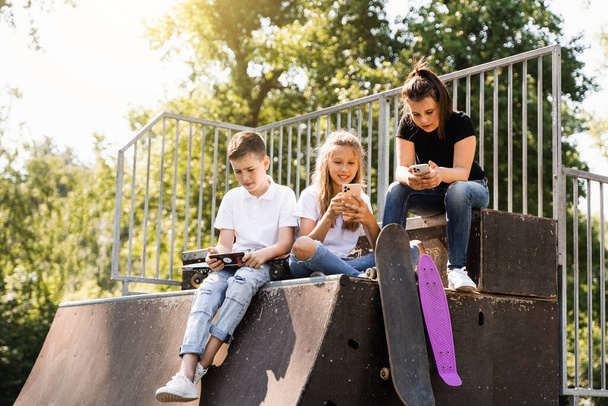 Зависимые от телефона спортивные дети со скейтбордом и пенни-бордами сидят и смотрят на смартфоны на спортивной рампе на детской площадке. Детская зависимость от телефонов - Фото, изображение