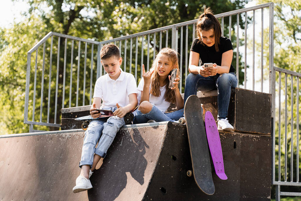 Phone addicted sports kids with skateboard and penny boards are sitting and looking at smartphones on sports ramp on playground. Children addiction of phones - Zdjęcie, obraz