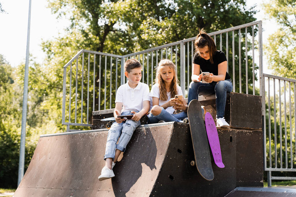Phone addicted sports kids with skateboard and penny boards are sitting and looking at smartphones on sports ramp on playground. Children addiction of phones - Photo, image