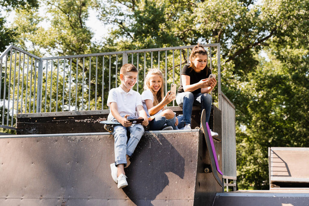 Phone addicted sports kids with skateboard and penny boards are sitting and looking at smartphones on sports ramp on playground. Children addiction of phones - Photo, Image