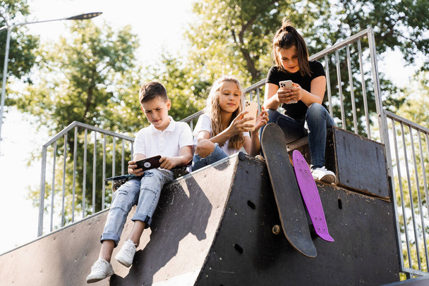 Phone addicted sports kids with skateboard and penny boards are sitting and looking at smartphones on sports ramp on playground. Children addiction of phones - Photo, image