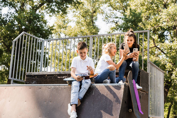 Phone addicted sports children with skateboard and penny boards are sitting and looking at smartphones on sports ramp on playground. Kids addiction of phones - Foto, Imagem