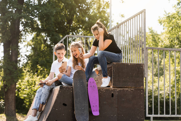 Phone addicted sports children with skateboard and penny boards are sitting and looking at smartphones on sports ramp on playground. Kids addiction of phones - Foto, imagen