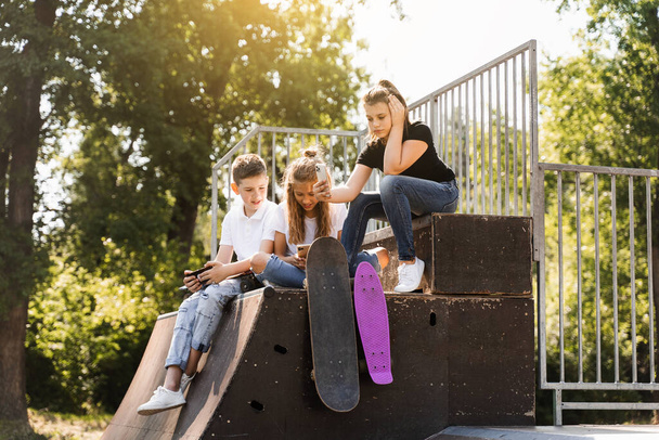 Phone addicted sports children with skateboard and penny boards are sitting and looking at smartphones on sports ramp on playground. Kids addiction of phones - Zdjęcie, obraz