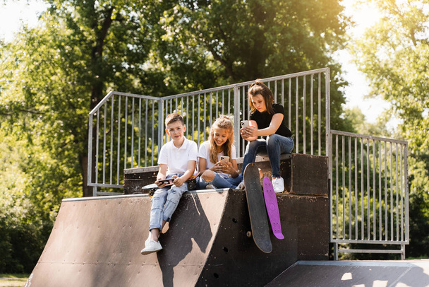 Τα εθισμένα στο τηλέφωνο παιδιά με skateboard και penny boards χρησιμοποιούν τηλέφωνα αντί για πατινάζ και παίζουν μαζί. Τα παιδιά κοιτάζουν smartphones στην αθλητική ράμπα. Παιδικός εθισμός στα τηλέφωνα - Φωτογραφία, εικόνα