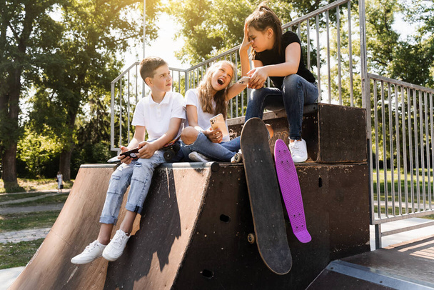Τηλέφωνο εθισμένα αθλητικά παιδιά με skateboard και penny boards χαμογελώντας και κοιτάζοντας smartphones σε αθλητικές ράμπα στην παιδική χαρά. Παιδικός εθισμός στα τηλέφωνα - Φωτογραφία, εικόνα