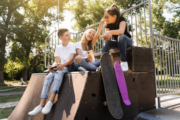 Phone addicted sports children with skateboard and penny boards smiling and looking at smartphones on sports ramp on playground. Kids addiction of phones - Photo, Image
