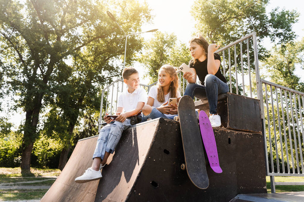 Τηλέφωνο εθισμένα αθλητικά παιδιά με skateboard και penny boards χαμογελώντας και κοιτάζοντας smartphones σε αθλητικές ράμπα στην παιδική χαρά. Παιδικός εθισμός στα τηλέφωνα - Φωτογραφία, εικόνα