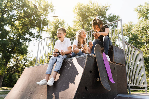 Phone addicted sports children with skateboard and penny boards smiling and looking at smartphones on sports ramp on playground. Kids addiction of phones - 写真・画像
