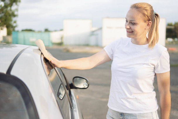 a girl of European appearance with blond hair in a white T-shirt, wipes the side windows in the car with a special yellow rag for windows. The concept of work and cleanliness. High quality photo - Фото, зображення