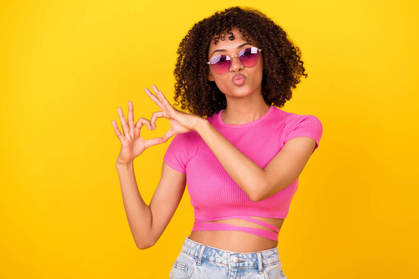 Photo of pretty shiny girl dressed pink t-shirt dark glasses sending kiss showing arms heart isolated yellow color background. - Fotó, kép
