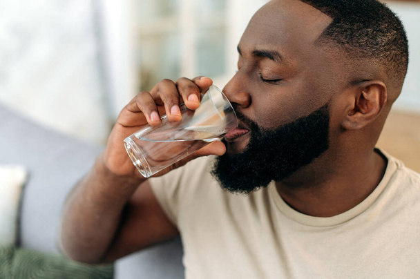 Leading a healthy lifestyle. Close-up of african american man, drinks a glass of clean water with pleasure, care his health, balances fluids in the body, replenishes energy and strength in the body - Foto, Bild