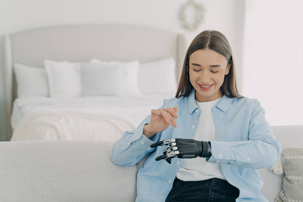 Happy girl is setting her electronic bionic limb. Attractive european woman with artificial arm at home. Smiling woman has modern cyber prosthesis. Engineering and innovation concept. - 写真・画像