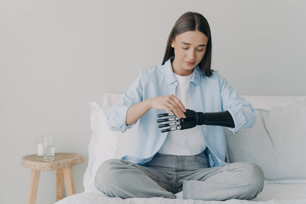 Disabled young woman is setting myoelectric prosthesis prosthesis parameters and functions. Bionic hand has processor chip and buttons. Attractive european girl adjusting her artificial arm at home. - 写真・画像