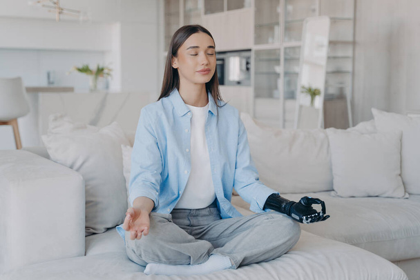 Tranquil disabled woman is sitting in lotus pose on couch with her eyes closed. Attractive caucasian girl is amputee. Lady has myoelectric cyber prosthesis. Modern artificial limb with sensor control. - Фото, изображение