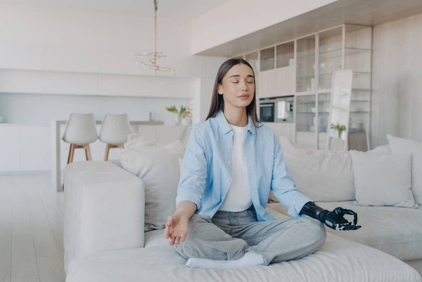 Morning yoga of disabled woman. Lady sitting in lotus pose on couch with her eyes closed. Attractive european girl has myoelectric cyber prosthesis. Modern artificial limb with nervous sensor control. - Foto, Imagem