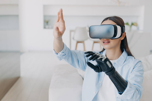 Handicapped girl with cyber bionic hand in virtual reality glasses at home. Disabled person gets rehabilitation. Young european woman sitting in living room on couch. Excited woman touches the vision. - Foto, imagen