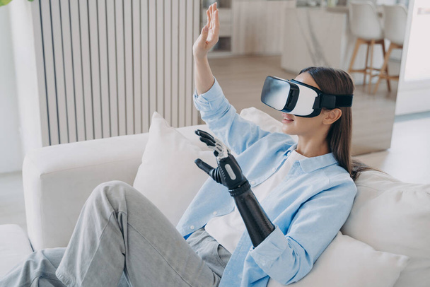 Handicapped girl in virtual reality glasses at home. Disabled person rehabilitation. European woman is getting used to modern bionic prosthesis using vr headset. Electronic technology in medicine. - Foto, immagini