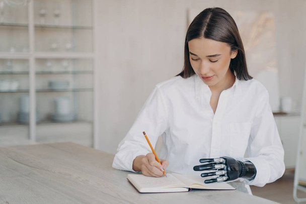 Disabled european girl is taking notes at the desk and doing homework. Happy handicapped young woman with futuristic cyber arm is studying remotely in college. Concept of equal opportunities. - Valokuva, kuva