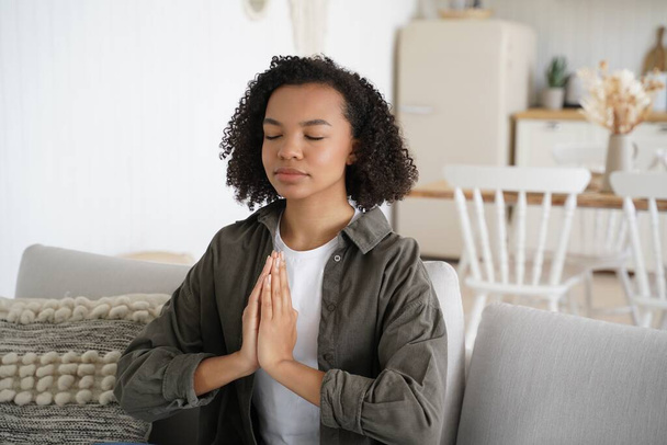 Teenage african american girl is practicing yoga at home. Curly young spanish woman is sitting on couch in lotus pose with her eyes closed. Harmony and balance. Wellness and spirituality. - Foto, Imagem
