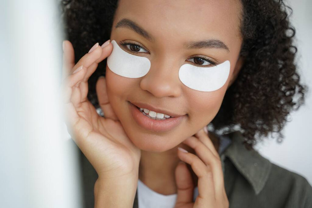 Young biracial girl with applied collagen patches on under eye skin smiling. Mixed race teen lady caring for under-eye skin, preventing puffiness, enjoying morning beauty routine. Skincare concept - Fotó, kép