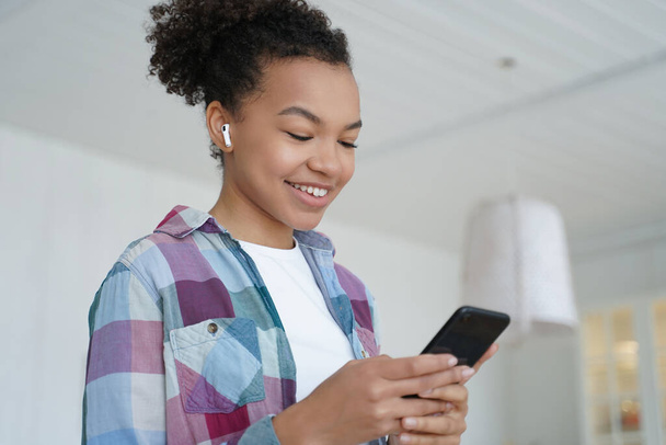 Music listening in airpods on mobile phone. Joyful teenage african american girl enjoying favourite song in wireless earphones. Student is browsing internet and social media on smartphone. - Photo, image
